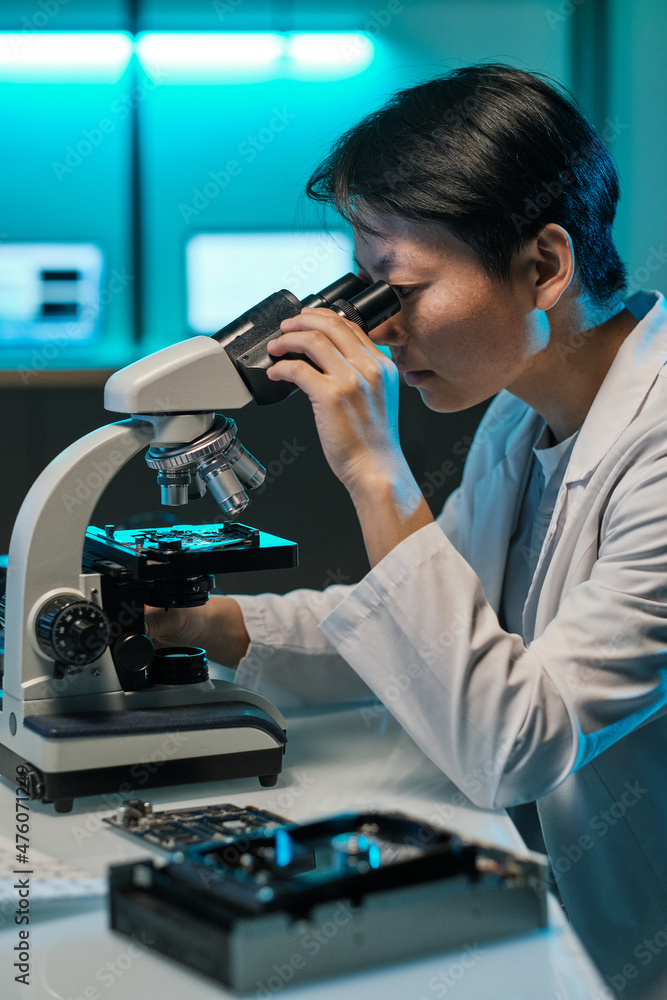 Poster Young Chinese woman in whitecoat looking in microscope lens while studying microchip scheme in laboratory