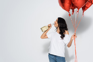 pretty brunette woman with closed eyes holding red balloons while drinking champagne from bottle on grey.