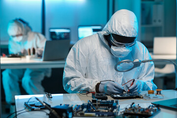 Technician in protective coveralls, eyeglasses, gloves and respirator repairing circuit board with...