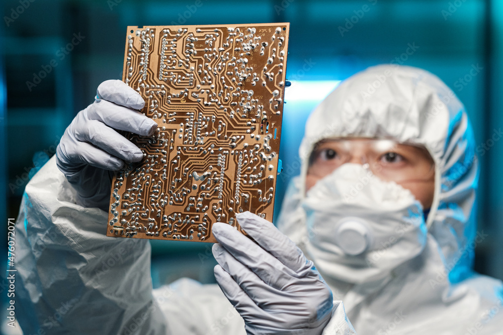 Poster gloved female specialist in protective coveralls and respirator holding circuit board in front of he