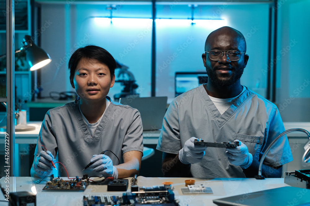 Poster Two young successful technicians in gloves and uniform working by desk and looking at camera in laboratory or workshop