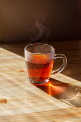 Glass cup of hot steaming tea under bright sunlight on a wooden table