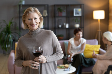 Portrait of mature woman holding glass of red wine and smiling at camera during a meeting with her friends