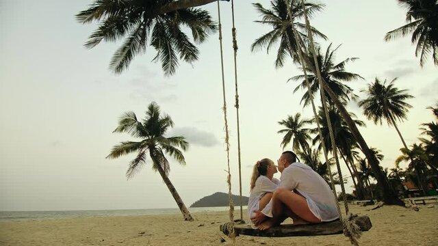 Loving couple on the swing on tropical beach. Woman and man in white shirt enjoy gold sunset and life in beaches, palm tree. Evening warm sunset