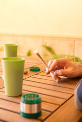 Man with a cannabis joint during a party in an outdoor and sunny place. Person at a table with grinder, marijuana and drinks, copy space top.
