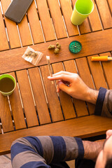 Young man sitting at a table with a marijuana joint, grinder and a drink in an outdoor place on sunny day.