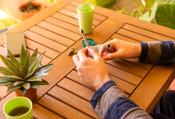 Unrecognized person at a wooden table smoking cannabis joint in a beautiful place full of plants.