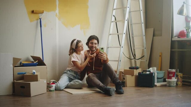Young couple man and woman taking photos selfie on smartphone while making overhaul. Husband and wife doing renovating together, painting walls in yellow. Interior design, new apartment. 