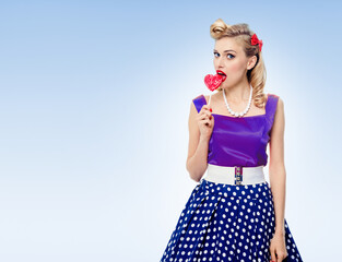 Portrait of woman eating heart shape lollipop dressed in pinup style dress in polka dot, over light blue background. Blonde pin up girl posing in retro fashion and vintage concept studio shot.