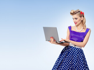 Portrait of beautiful woman holding laptop, dressed in pin-up style dress in polka dot, over light blue background. Blonde pinup girl posing in retro fashion and vintage concept studio shot.