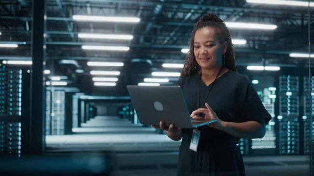 Portrait of Successful African American Female IT Specialist Using Laptop, Standing in Data Center. System Administrator Works on Web Services. Cloud Computing, Server Analytics, Cyber Security