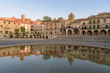 Naklejka na ściany i meble City landscape of the main square of the medieval city of Trujillo with its old stone buildings and arcades reflected in the water of the fountain at sunrise, Caceres, Extremadura, Spain