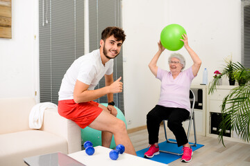 senior elderly woman with a personal trainer private fitness coach at home handsome young man