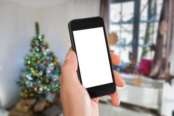 Hand of human holding the phone with empty screen mockup near decorated festive tree, celebrating Christmas,