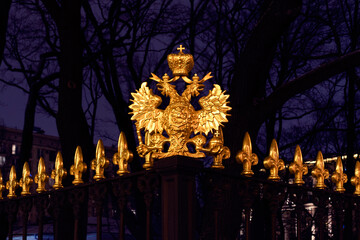 Gilded coat of arms of Russia, two-headed eagle on fence at night against background of trees