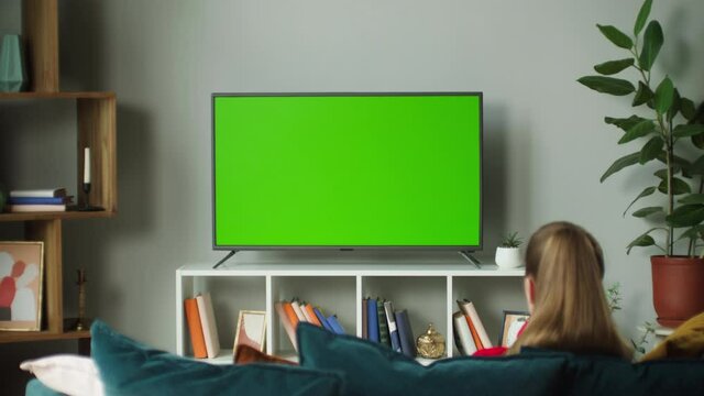 Woman watching television with green screen. Unrecognizable female person switching channels on tv with chroma key, sitting on sofa in living room. Spare time at home, relaxing and leisure.