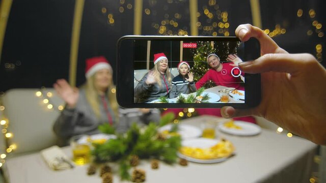 Close-up of the phone in the hands of a girl taking pictures of a family with a child for the New Year. Happy family waving his hands at camera during christmas party at