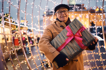 Portrait of senior man with wrapped present at Christmas market during winter evening. - obrazy, fototapety, plakaty