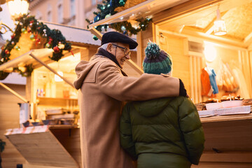 Embraced grandfather and grandson buying street food at Christmas market stall.