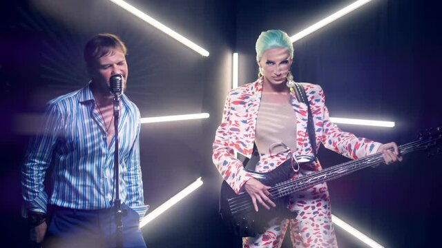 travesty actor plays electric guitar on stage and handsome man sings into microphone.