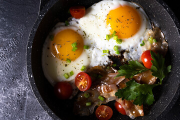 breakfast with fried eggs in a pan with bacon and vegetables on dark background
