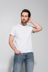 young man in white t-shirt holding hand in pocket of jeans, touching hair and looking away on grey.