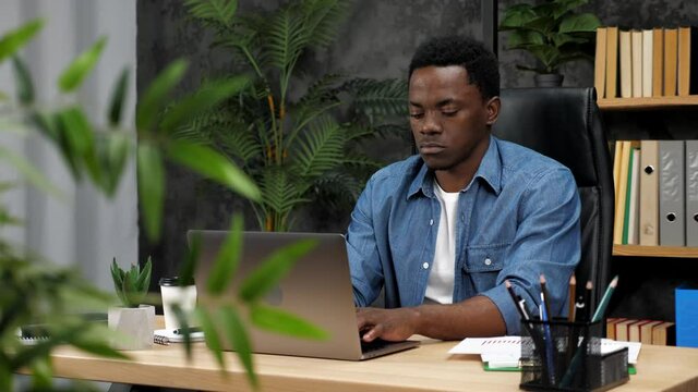 Confident adult African American man freelancer works for laptop typing on keyboard. Male stock trader businessman uses computer chatting with company employees, sitting on chair at desk in office