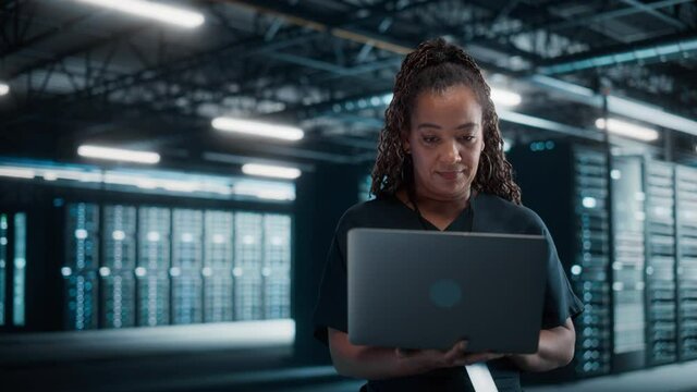 African American Female IT Specialist Using Laptop Computer, Standing in Big Warehouse Data Center. System Administrator working with SAAS, Cloud Web Services. e-Business Entrepreneur. Static