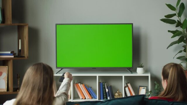 Women watching television with chroma green screen. Girlfriends switching channels on tv with remote controller, sitting on sofa together in living room. Spare time at home, relaxing.