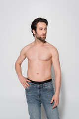 shirtless man in jeans looking away while standing with hand on hip on grey.