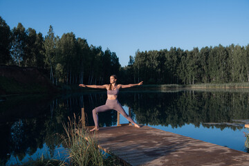 Fototapeta na wymiar Young woman on wooden pier above forest lake scenery, folds her arms in a namaste gesture. Woman arms outstretched in nature.