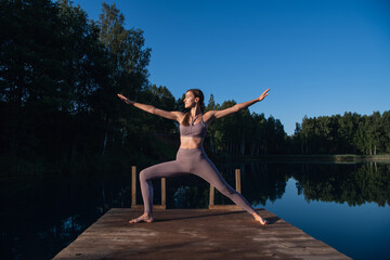 Fototapeta na wymiar Young woman on wooden pier above forest lake scenery, folds her arms in a namaste gesture. Woman arms outstretched in nature.