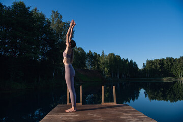 Fototapeta na wymiar Young female practicing yoga against the sky on a sunrise. Pretty slim woman performs an exercise. Healthcare, meditaion, welness, mindfulness concept.