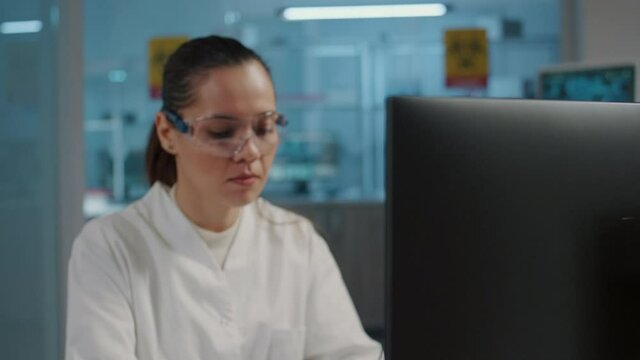 Woman chemist working on computer in science laboratory, using technology to develop microbiology experiment with lab equipment. Scientist with protective glasses. Handheld shot