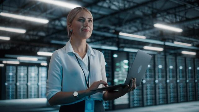 Successful Female Chief Technology Officer Using Laptop Computer, Standing in Big Warehouse Data Center. System Administrator Maintenance of SAAS, Cloud Services Server. Digital Entrepreneur. Static
