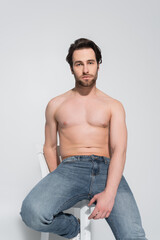 brunette shirtless man in jeans sitting on white chair and looking at camera on grey.