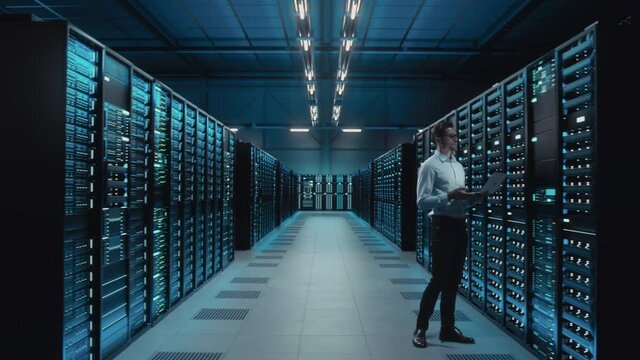 Big Data Center IT Specialist Using Laptop Computer. Server Farm Cloud Computing Facility with System Administrator Working. Software as Solution, Cyber Security. Moving Between Rack Raws Dolly Shot