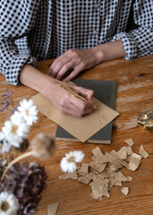 Person in a plaid shirt sitting behind a wooden table and writing on a piece of craft paper. Dry flowers and scraps of paper complete the composition.