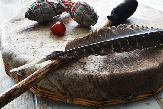 A side view image of a hand made leather meditation drum with sacred feather and two white sage smudge sticks. 