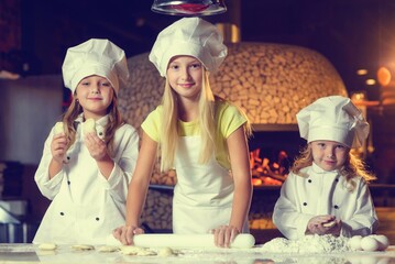 Near the oven, three cheerful little girls, soiled in flour and dressed in chef's uniforms, are making pizza.