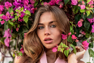 Fototapeta premium Beauty portrait of natural young attractive woman with freckles on her face posing in pink bush of flowers.