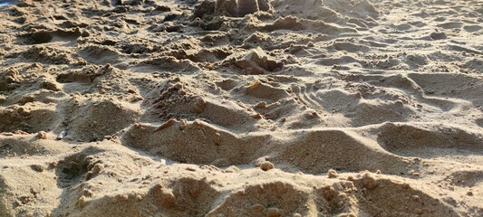 Beach sand in the rays of the summer sun. Sunny day on the shore. The yellow-brown sand is uneven with dents and footprints of people. Bumps in the sand cast dark shadows from the sun.