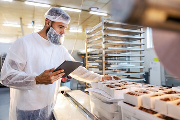 A food inspector in a sterile white uniform is holding the tablet and looking at collected cookies....