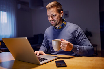 Remote business from home. A young smiling businessman with earphones sitting at home, typing on a laptop and holding a mug with coffee. A man having an online meeting.