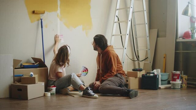 Young couple making overhaul, sitting on floor and choosing colors, renovation, painting walls together. Man and woman talking about changing interior design, new apartment.