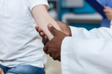 Closeup of african american pediatrician doctor rubbing injured arm of little patient during medical physiotherapy in hospital office. Young kid having broken bone after accident. Health care service