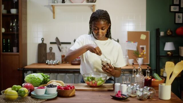 African american chef cooking vegetable salad. Woman adding spices and trying food, mixing dish. Happy housewife in cozy kitchen. Healthy vegetarian meal. 