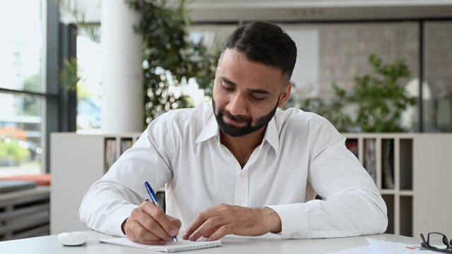 Online lecture. Friendly focused confident young adult Indian businessman, manager with a beard, wearing white shirt, sit at a table in modern office, taking notes during video conference, smiling