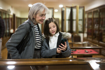 Cute curious tween girl browsing internet on smartphone in search of information about showpieces while visiting local history museum with her grandmother