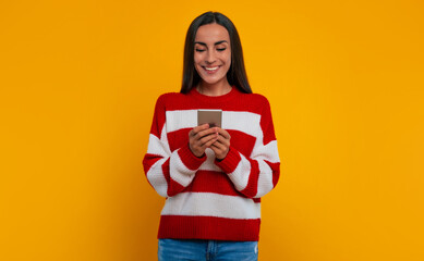 Beautiful young happy woman in a sweater while she using smart phone and isolated on yellow background